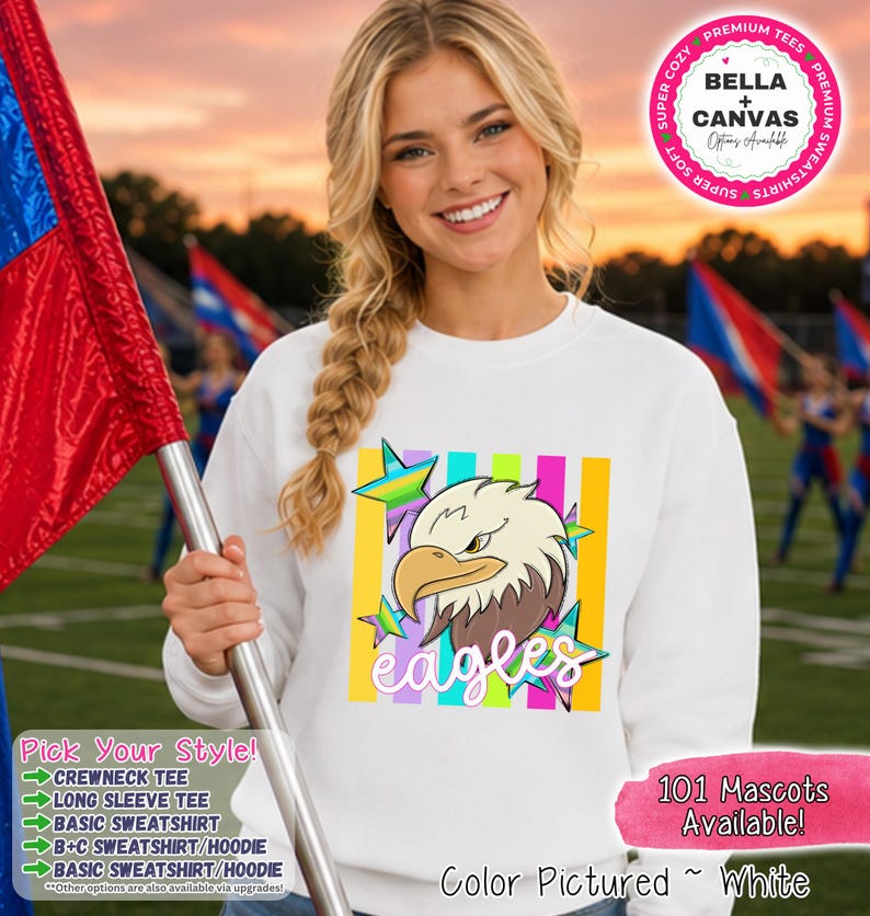 May include: A white crewneck sweatshirt featuring a graphic of a bald eagle head with a yellow beak and brown feathers. The design includes colorful vertical stripes in the background and multi-colored stars, with the word eagles in white script.
