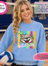 May include: A light blue crewneck sweatshirt featuring a graphic of a snarling wildcat head centered over vertical neon stripes and stars. The word wildcats is printed in white script below the cat. The sweatshirt is shown with a volleyball.