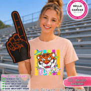 May include: A peach-colored crewneck t-shirt featuring a graphic of a tiger face surrounded by colorful stars and vertical stripes with the word tigers written below. The shirt is displayed with a black foam finger with orange trim and text.