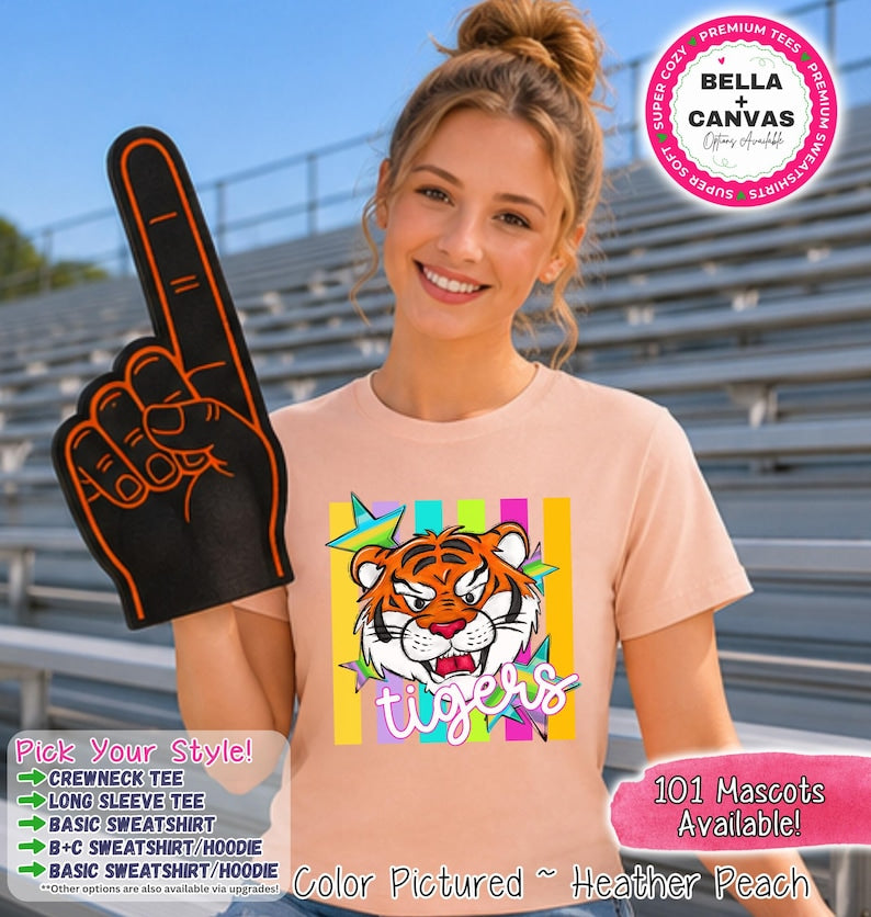 May include: A peach-colored crewneck t-shirt featuring a graphic of a tiger face surrounded by colorful stars and vertical stripes with the word tigers written below. The shirt is displayed with a black foam finger with orange trim and text.