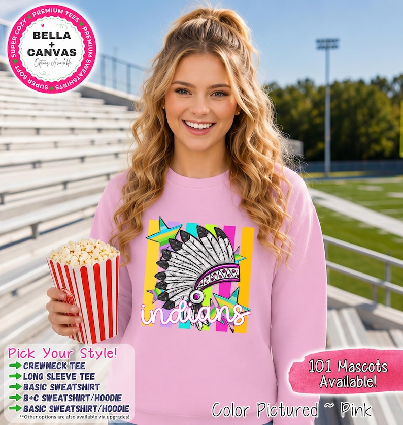 May include: A light pink crewneck sweatshirt featuring a graphic print of a feathered headdress in black, white, and purple, layered over colorful vertical stripes and stars, with the word indians written in white script lettering across the front.