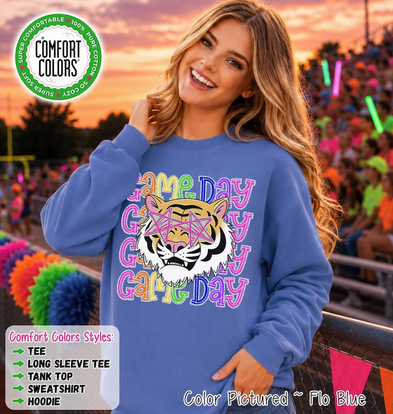 May include: A periwinkle blue crewneck sweatshirt featuring a graphic of a tiger wearing pink star-shaped glasses. The text "GAME DAY" is repeated in a colorful, stitched-style font behind the tiger. The sweatshirt has long sleeves.