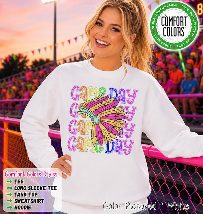 May include: A white long-sleeve sweatshirt featuring the repeated text "Game Day" in colorful, patterned lettering. Centered over the text is a graphic of a feathered headdress in shades of pink, yellow, and teal. A circular logo in the top right corner reads "Comfort Colors" with "Super Comfortable," "100% Pure Cotton," and "So Cozy" text.