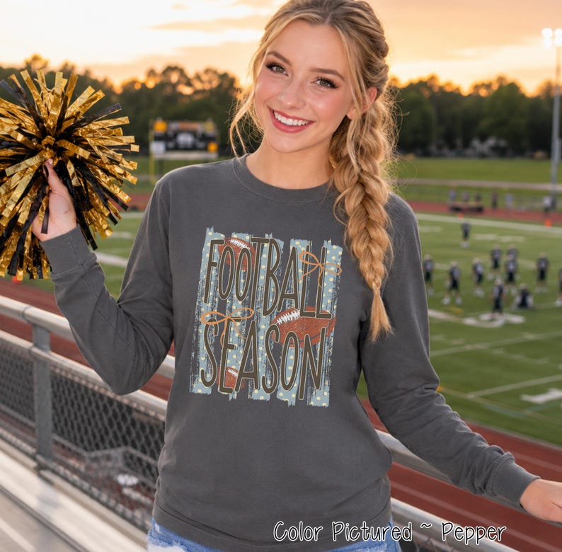 May include: A charcoal gray long-sleeve shirt featuring a graphic print with the text FOOTBALL SEASON in dark brown letters. The design includes light blue vertical brush strokes, white dots, and brown football illustrations on the front.
