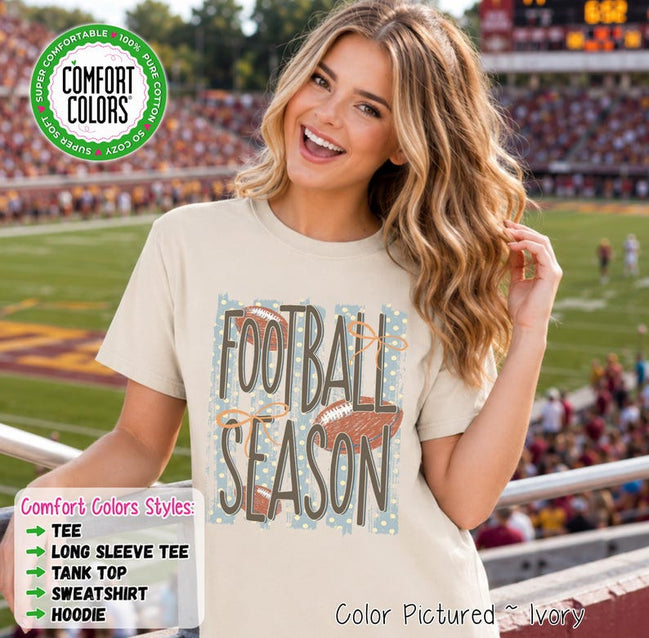 May include: An ivory short-sleeve t-shirt featuring a graphic print with the text "FOOTBALL SEASON" in dark brown letters. The design includes illustrations of three brown footballs and light blue vertical stripes with white polka dots.