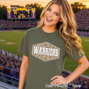 An olive green short-sleeved t-shirt featuring a central hexagonal graphic. The graphic contains a brown and tan camouflage pattern with the word WARRIORS printed in bold white capital letters across the center of the shape.