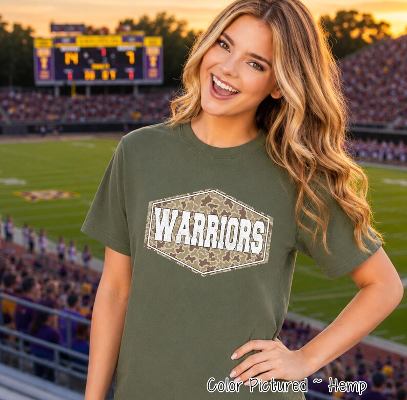 An olive green short-sleeved t-shirt featuring a central hexagonal graphic. The graphic contains a brown and tan camouflage pattern with the word WARRIORS printed in bold white capital letters across the center of the shape.