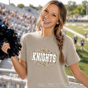 A sandstone-colored short-sleeve t-shirt featuring a central hexagonal graphic with a camouflage pattern and the word KNIGHTS printed in white block letters. The shirt is held alongside a black pom-pom in a stadium setting.