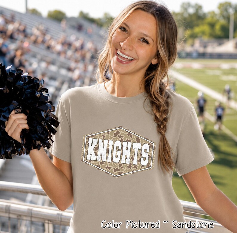 A sandstone-colored short-sleeve t-shirt featuring a central hexagonal graphic with a camouflage pattern and the word KNIGHTS printed in white block letters. The shirt is held alongside a black pom-pom in a stadium setting.