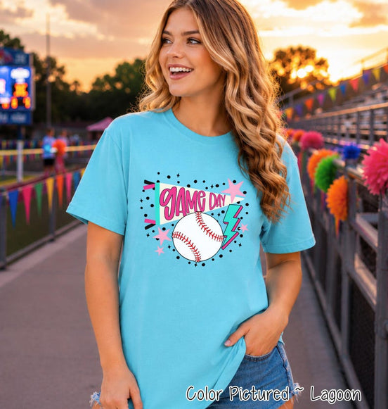 A lagoon blue short-sleeve t-shirt featuring a graphic print with the text "GAME DAY" in pink, a white baseball with red stitching, a pink star, and a teal lightning bolt, surrounded by small black dots and star accents.