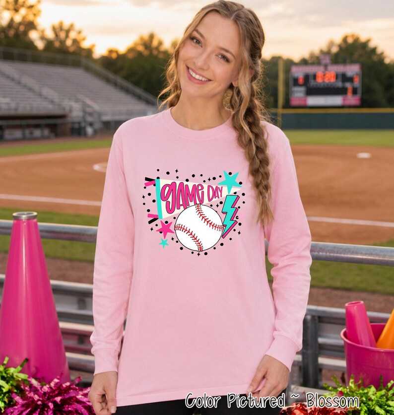A light pink long-sleeve shirt featuring a graphic print on the chest. The design includes a white baseball with red stitching, a teal lightning bolt, pink stars, and a teal pennant flag with the text 