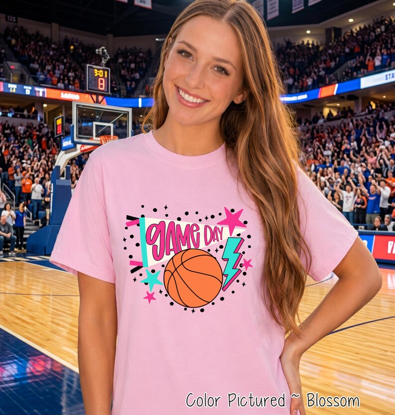 A light pink short-sleeve t-shirt featuring a graphic print with the text GAME DAY in pink letters, an orange basketball, a teal lightning bolt, a white megaphone, and various pink and teal stars surrounded by black polka dots.
