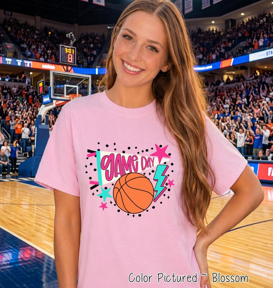 A light pink short-sleeve t-shirt featuring a graphic print with the text GAME DAY in pink letters, an orange basketball, a teal lightning bolt, a white megaphone, and various pink and teal stars surrounded by black polka dots.