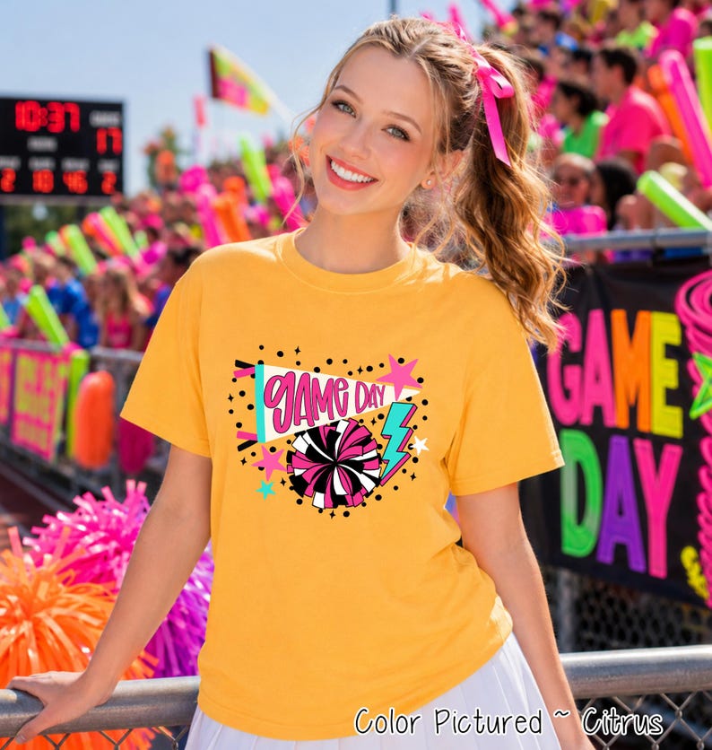 A citrus yellow short-sleeve t-shirt featuring a graphic print with the text "GAME DAY" in pink script, a megaphone, a black and pink pom-pom, a blue lightning bolt, and small pink stars against a background of black dots.