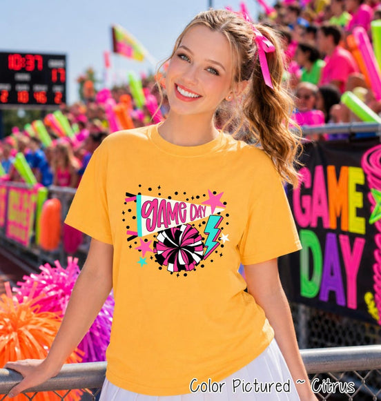 A citrus yellow short-sleeve t-shirt featuring a graphic print with the text "GAME DAY" in pink script, a megaphone, a black and pink pom-pom, a blue lightning bolt, and small pink stars against a background of black dots.