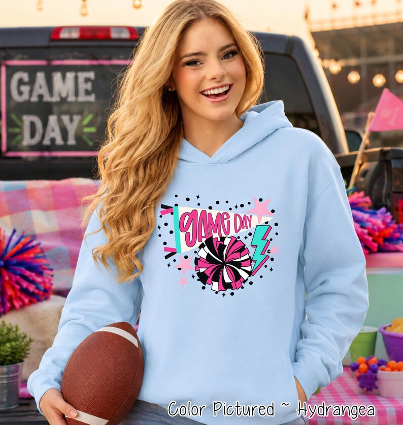 A light blue hooded sweatshirt featuring a graphic print with the text "GAME DAY" in pink, accompanied by a megaphone, a black and pink pom-pom, a teal lightning bolt, and pink stars, all set against a background of black dots.