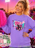 A violet long-sleeve sweatshirt featuring a graphic print with the text GAME DAY, a megaphone, a pom-pom, stars, and a lightning bolt. The design includes pink, white, and teal accents. A bright pink pom-pom is held to the side.