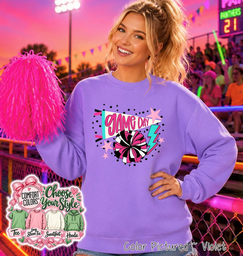 A violet long-sleeve sweatshirt featuring a graphic print with the text GAME DAY, a megaphone, a pom-pom, stars, and a lightning bolt. The design includes pink, white, and teal accents. A bright pink pom-pom is held to the side.
