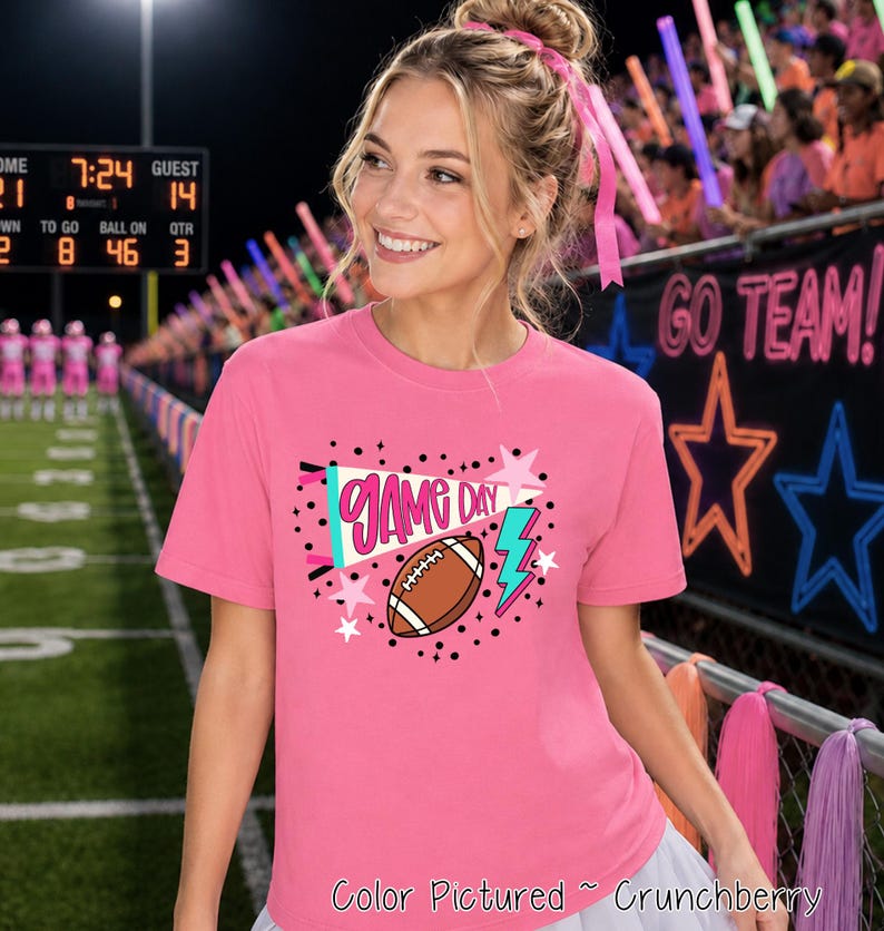 A bright pink short-sleeved t-shirt featuring a graphic print with the text 'GAME DAY' in pink, a brown football, a teal megaphone, a teal lightning bolt, and various pink and white stars surrounded by small black dots.
