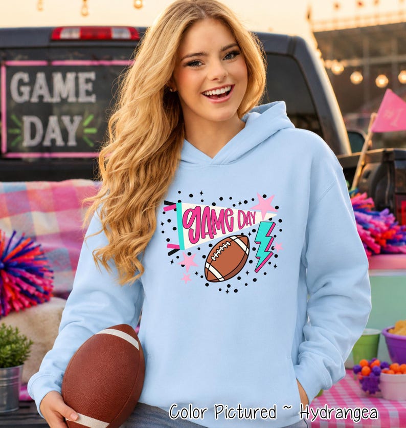 A light blue hooded sweatshirt featuring a graphic print on the chest with the text GAME DAY, a brown football, a pink megaphone, a pink star, and a teal lightning bolt. The hoodie is made of a soft fabric and has long sleeves.