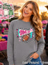 A granite gray long-sleeve shirt featuring a graphic print with the text GAME DAY in pink, a brown football, a teal and white pennant, a teal lightning bolt, and pink stars, surrounded by black dots on a gray fabric background.
