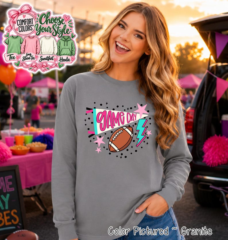 A granite gray long-sleeve shirt featuring a graphic print with the text GAME DAY in pink, a brown football, a teal and white pennant, a teal lightning bolt, and pink stars, surrounded by black dots on a gray fabric background.