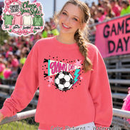 A watermelon-colored crewneck sweatshirt featuring a graphic print with the text GAME DAY in pink, a black and white soccer ball, a teal lightning bolt, and pink stars. The design is centered on the front of the long-sleeved garment.