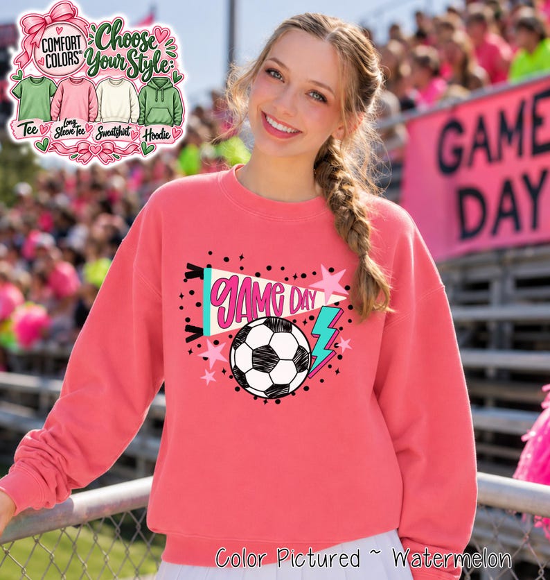A watermelon-colored crewneck sweatshirt featuring a graphic print with the text GAME DAY in pink, a black and white soccer ball, a teal lightning bolt, and pink stars. The design is centered on the front of the long-sleeved garment.