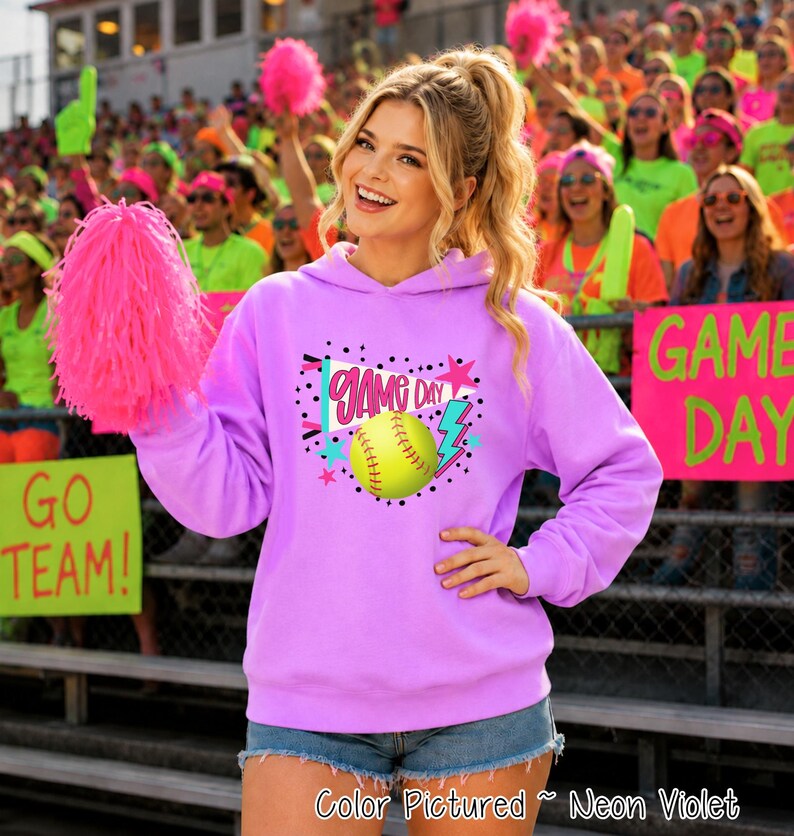 A neon violet hooded sweatshirt featuring a graphic print on the chest. The design includes a yellow softball, a blue megaphone with the text GAME DAY, a pink star, and a blue lightning bolt, all surrounded by black polka dots.