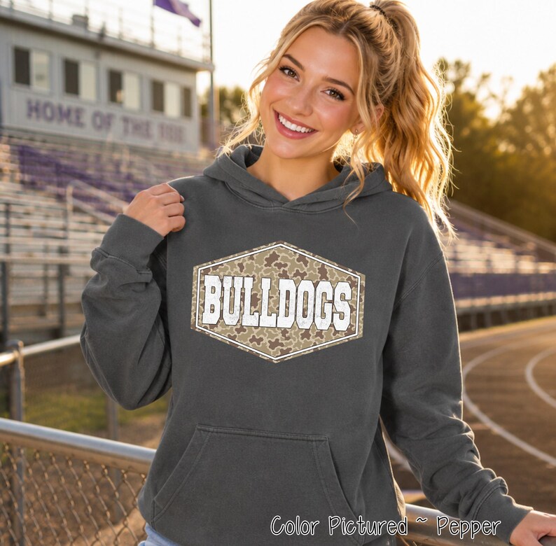 A pepper-colored hooded sweatshirt featuring a central hexagonal graphic with a camouflage pattern and the white text BULLDOGS in a bold, block-style font. The garment includes a front kangaroo pocket and long sleeves.