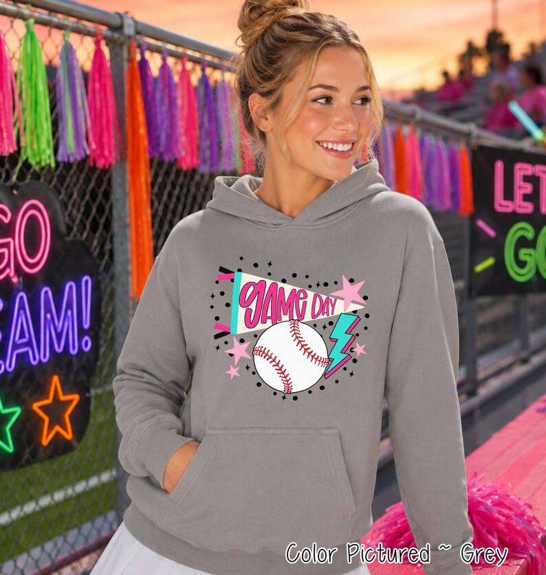 A grey hooded sweatshirt featuring a graphic print on the front. The design includes a white baseball with red stitching, a pink pennant flag with the text GAME DAY, a blue lightning bolt, and several small pink stars and black dots.