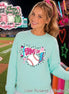 A light mint green crewneck sweatshirt featuring a graphic print on the chest. The design includes a white pennant flag with the text GAME DAY in pink, a white baseball with red stitching, a blue lightning bolt, and pink stars.