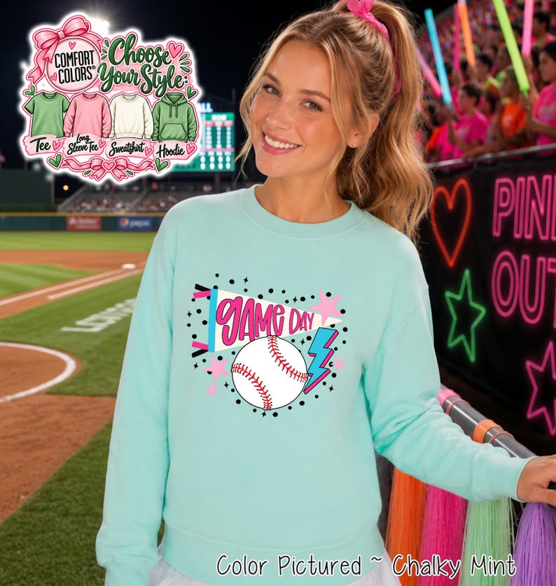 A light mint green crewneck sweatshirt featuring a graphic print on the chest. The design includes a white pennant flag with the text GAME DAY in pink, a white baseball with red stitching, a blue lightning bolt, and pink stars.