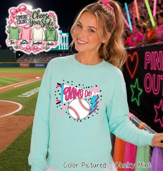 A light mint green crewneck sweatshirt featuring a graphic print on the chest. The design includes a white pennant flag with the text GAME DAY in pink, a white baseball with red stitching, a blue lightning bolt, and pink stars.