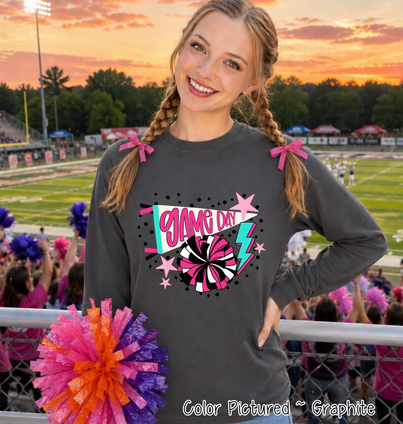A charcoal gray long-sleeve crewneck sweatshirt featuring a graphic print with the text "GAME DAY" in pink, a pink and black pom-pom illustration, a teal pennant, and pink stars and lightning bolts on the front chest.