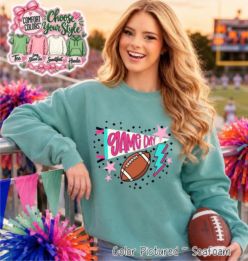 A seafoam green crewneck sweatshirt featuring a graphic print with the text GAME DAY in pink, a brown football, a light blue lightning bolt, and pink stars. A Comfort Colors chart shows options for a tee, long sleeve, sweatshirt, and hoodie.