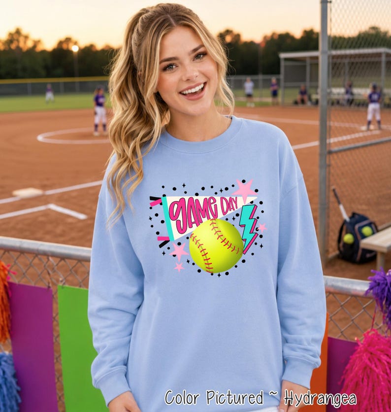 A light blue crewneck sweatshirt featuring a graphic print on the chest. The design includes a yellow softball, a white pennant with the text "GAME DAY" in pink, a blue lightning bolt, and pink stars surrounded by black dots.