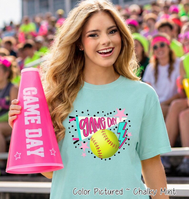 A chalky mint short-sleeve t-shirt featuring a graphic print of a yellow softball, a pink pennant with the text GAME DAY, a pink lightning bolt, and pink stars. A bright pink megaphone with the white text GAME DAY is also shown.