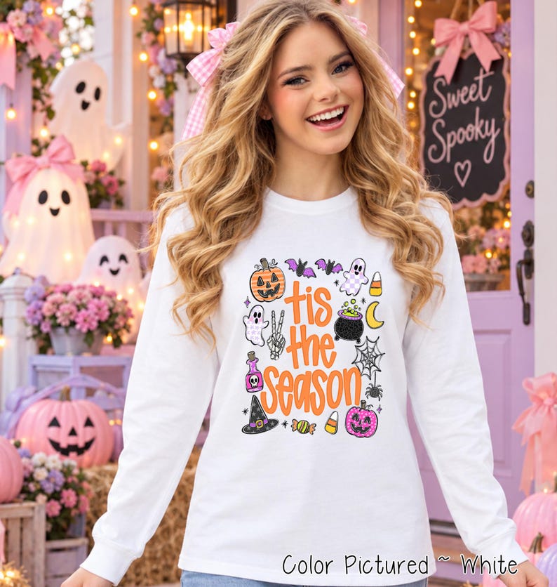 A white long-sleeve shirt featuring a colorful graphic print with the text 'tis the season' in orange. The design includes illustrations of a jack-o'-lantern, ghosts, a cauldron, a witch hat, a spider web, bats, and candy corn.
