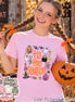 A light pink t-shirt featuring a graphic print with the text 'tis the season' in orange, surrounded by Halloween-themed illustrations including a pumpkin, ghosts, a cauldron, a spider web, a witch hat, and candy corn.