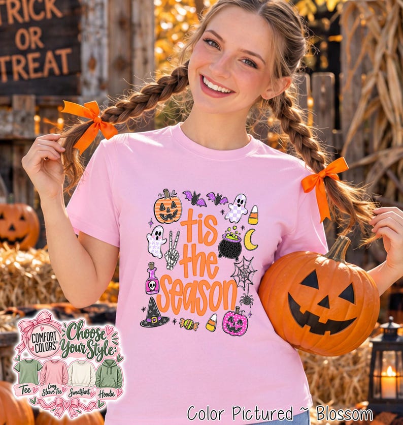 A light pink t-shirt featuring a graphic print with the text 'tis the season' in orange, surrounded by Halloween-themed illustrations including a pumpkin, ghosts, a cauldron, a spider web, a witch hat, and candy corn.