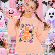 A peach-colored crewneck sweatshirt featuring a graphic print with the text 'tis the season' in orange. The design includes illustrations of a jack-o'-lantern, ghosts, a cauldron, a spider web, a witch hat, candy corn, and bats.
