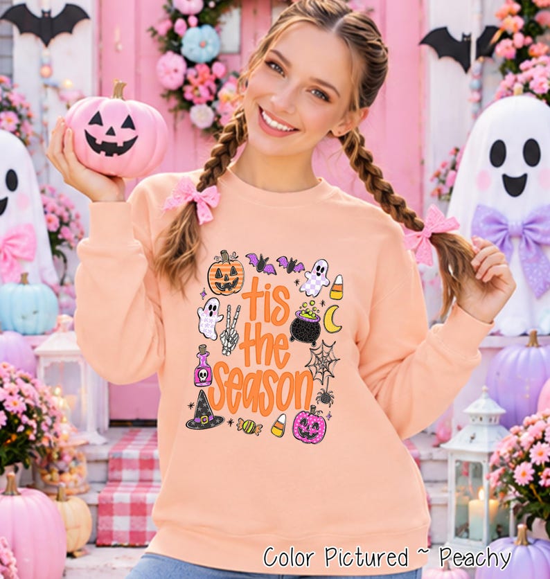 A peach-colored crewneck sweatshirt featuring a graphic print with the text 'tis the season' in orange. The design includes illustrations of a jack-o'-lantern, ghosts, a cauldron, a spider web, a witch hat, candy corn, and bats.