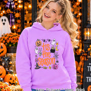 A neon violet hooded sweatshirt featuring a graphic print with the text 'tis the season' in orange. The design includes illustrations of a jack-o'-lantern, ghost, bat, candy corn, cauldron, spider web, potion bottle, and witch hat.