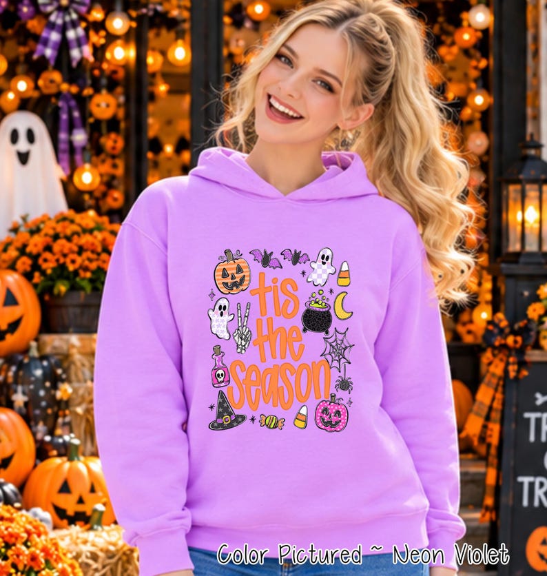 A neon violet hooded sweatshirt featuring a graphic print with the text 'tis the season' in orange. The design includes illustrations of a jack-o'-lantern, ghost, bat, candy corn, cauldron, spider web, potion bottle, and witch hat.